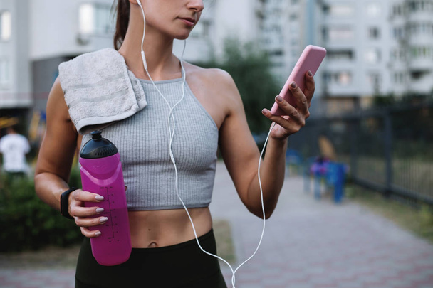 Girl athlete in a break between training chooses the music for jogging on the phone holding a bottle of water. - Foto, Imagen