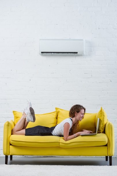 happy young woman working with laptop while lying on couch under air conditioner hanging on wall - Фото, изображение