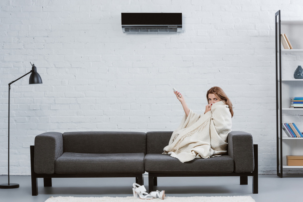 νεαρή γυναίκα που καλύπτεται με κουβέρτα καναπέ κάτω από το κλιματιστικό που κρέμονται στον τοίχο - Φωτογραφία, εικόνα