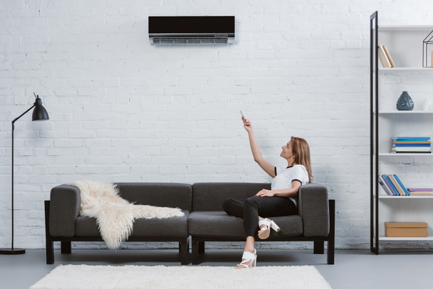 bella giovane donna seduta sul divano e che punta al condizionatore d'aria appeso al muro con telecomando
 - Foto, immagini