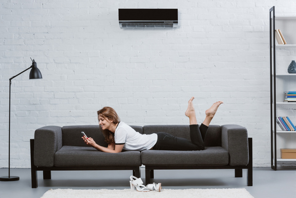 heureuse jeune femme en utilisant smartphone tout en étant couché sur le canapé sous climatiseur accroché au mur
 - Photo, image