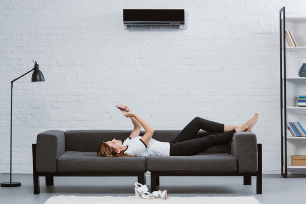 feliz joven mujer usando teléfono inteligente en el sofá bajo el aire acondicionado colgando en la pared
 - Foto, imagen