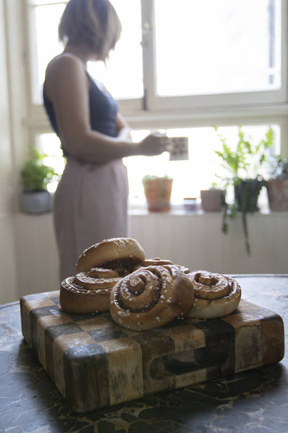 Freshly baked cinnamon buns stacked in a pile with a woman drinking coffee in the background - Photo, Image