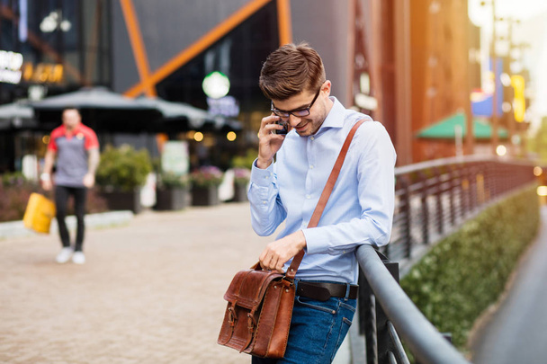 waiting for the coffee meeting. handsome smart casual man with eyeglasses and leather bag standing outside against a metal fence in front of comercial center talking at mobile phone - Photo, image