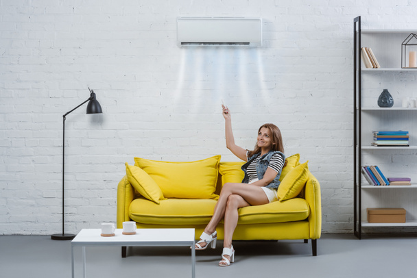 aantrekkelijke jonge vrouw wijzend op de airconditioner opknoping op een muur met afstandsbediening - Foto, afbeelding