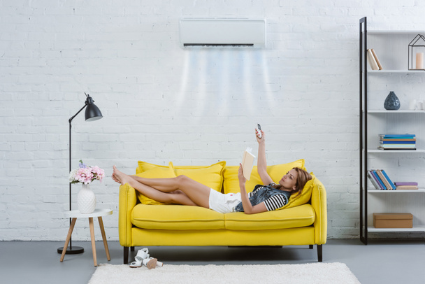concentrated young woman reading book on couch and pointing at air conditioner hanging on wall with remote control - Fotoğraf, Görsel