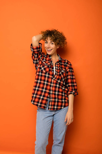 excited curly woman in a checkered shirt and jeans standing over an orange background with empty space for text next to her - Zdjęcie, obraz