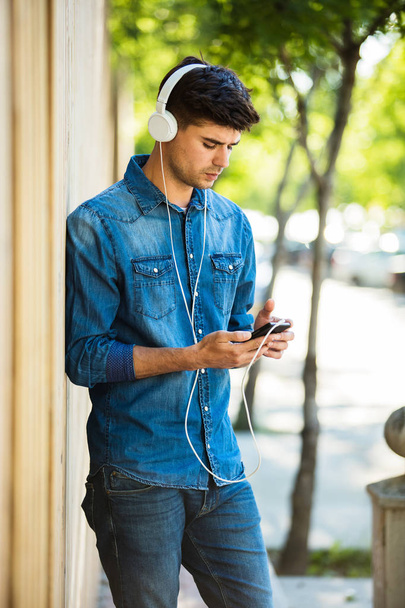 modern and stylish man listening music on headsets outside on the street on a sunny day, checking his phone - Foto, Imagen