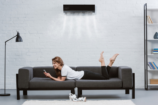 счастливая молодая женщина с помощью смартфона на диване под кондиционером висит на стене
 - Фото, изображение