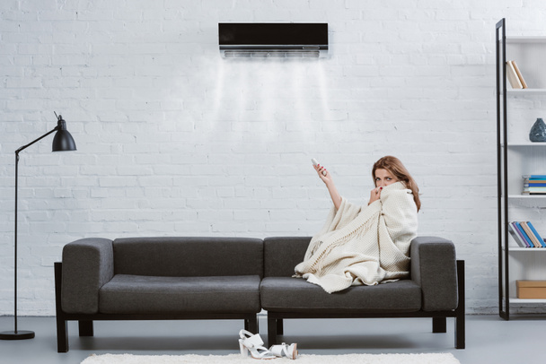 jovem coberta com cobertor no sofá sob ar condicionado pendurado na parede e soprando ar refrigerado
 - Foto, Imagem