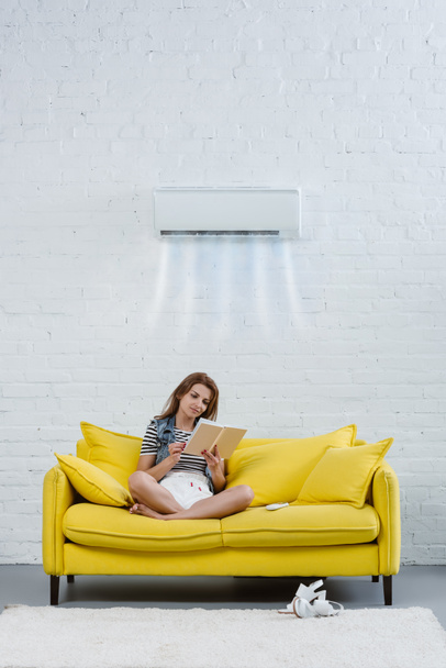 エアコンが壁に掛かっている冷却空気を吹き下のソファで本を読む若い女性に焦点を当てた - 写真・画像