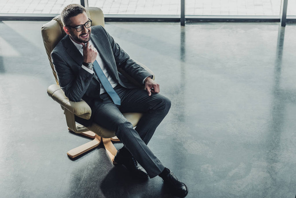 hoge hoekmening van knappe gelukkig zakenman zittend op luxe fauteuil op moderne kantoor - Foto, afbeelding