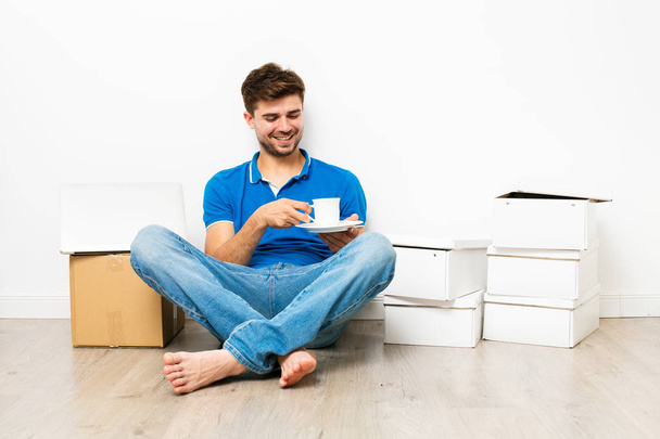 young man in blue shirt having a coffee or tea on the floor next to his cardbox, mooving to new house. on white background - Valokuva, kuva