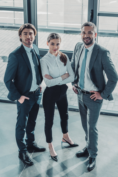Skupina úspěšných mladých podnikatelů stojící v moderní kanceláři a při pohledu na fotoaparát - Fotografie, Obrázek