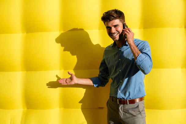 elegant smart casual guy with cellphone presenting an offer on his side, standing on a yellow background - Φωτογραφία, εικόνα