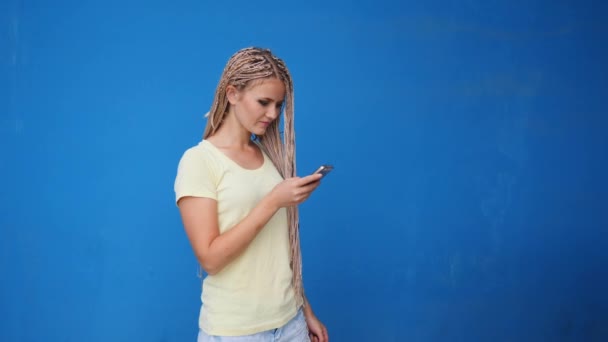 Smartphone woman talking on phone laughing over blue background. Beautiful young female with braids having casual conversation on mobile phone - Video