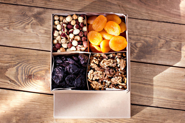Ένα κουτί με ξηρούς καρπούς και αποξηραμένα φρούτα σε παλιό ξύλινο φόντο... Αποξηραμένα βερίκοκα, δαμάσκηνα, καρύδια, φουντούκια. - Φωτογραφία, εικόνα