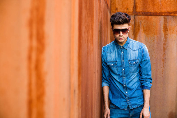 young sexy man in jeans outfit and sunglasses standing against a rusty wall - Photo, image