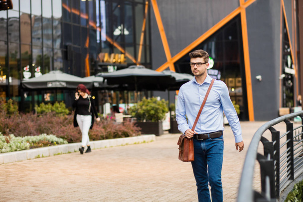 successfull entrepreneur. handsome smart casual man with eyeglasses and leather bag walking outside with confidence and smile - Fotó, kép