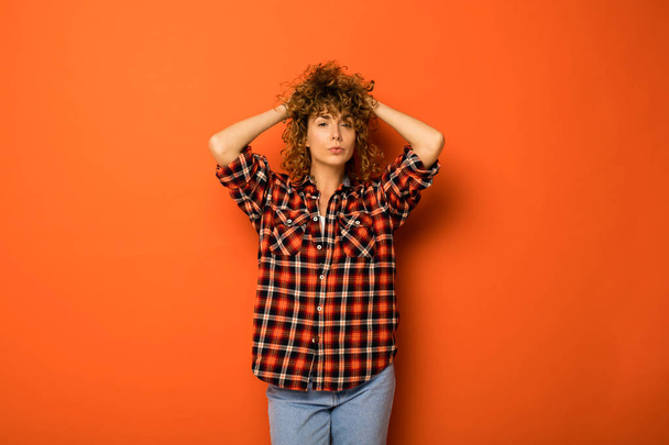 pretty curly woman in a checkered shirt and jeans standing over an orange background with empty space for text next to her holding hands on the head - Foto, imagen