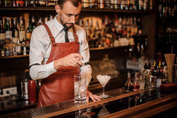 Barman na camisa branca e avental de couro marrom mexendo um cubo de gelo no copo de coquetel no balcão do bar
 - Foto, Imagem