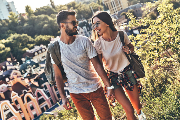 smiling Caucasian couple in casual clothing walking outdoors, city houses on background  - Фото, изображение