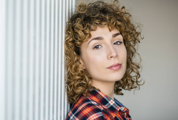 young natural curly woman smiling to camera casual dressed in a checkered shirt inside a house - Foto, Imagem