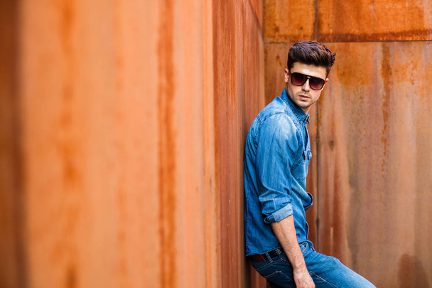 young sexy man in jeans outfit and sunglasses standing against a rusty wall - Φωτογραφία, εικόνα