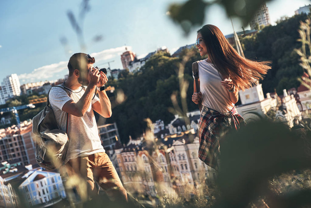 man fotograferen zijn gelukkig vriendin terwijl staande op de heuvel, de huizen van de stad op de achtergrond  - Foto, afbeelding