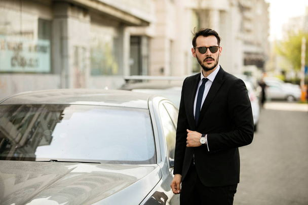eleganter Mann im Anzug, erfolgreicher Geschäftsmann, der neben seiner Limousine steht und selbstbewusst aussieht - Foto, Bild