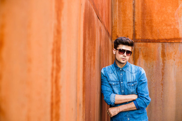 young sexy man in jeans outfit and sunglasses standing against a rusty wall - Zdjęcie, obraz