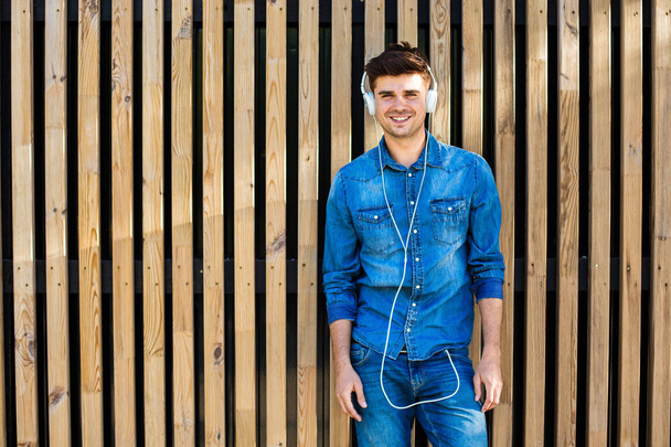 cool and modern. handsome sexy man in jeans outfit, on wooden background, with free space for text next to him, standing confident and cheerful, relaxing with music in his ear - Photo, image