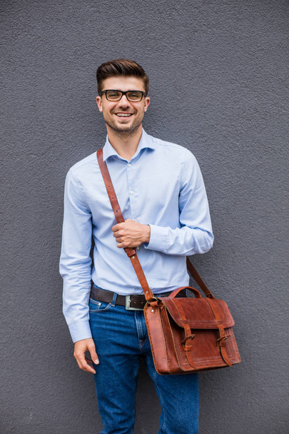 smart and cool. smart casual man with eyeglasses and leather bag standing on gray background, wall, confident and relaxed - 写真・画像