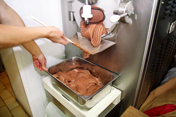 Confectioner in chef uniform is working on ice cream maker machine. Producing black chocolate ice cream flavors and it falls into steel container. Industrial preparation of italian creamy ice cream. - Photo, image