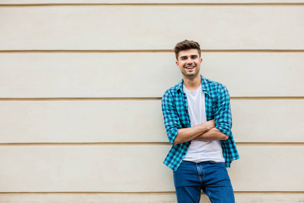 handsome man in checkered shirt smiling standing outside leaning on a wall with free space next to him - Фото, изображение
