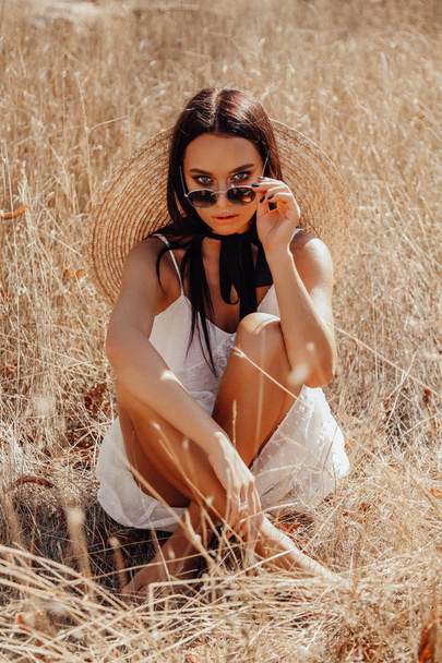 fashion summer outdoor photo of beautiful sexy woman with dark hair in elegant dress with luxurious straw hat and bag posing in field with dry grass - Photo, image