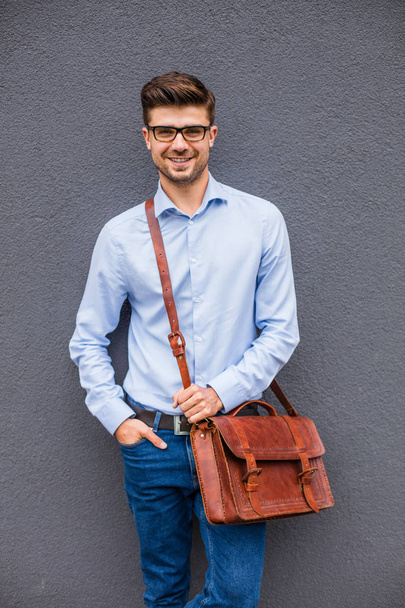 smart and cool. smart casual man with eyeglasses and leather bag standing on gray background, wall, confident and relaxed - Photo, image