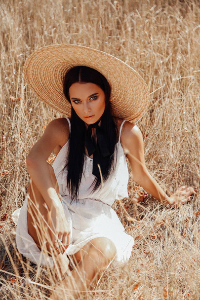 fashion summer outdoor photo of beautiful sexy woman with dark hair in elegant dress with luxurious straw hat and bag posing in field with dry grass - Photo, Image