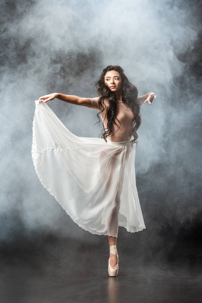 beautiful young ballerina in white skirt dancing on dark background with smoke around - Photo, Image