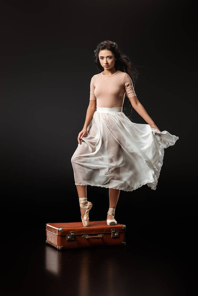 elegant ballerina in white skirt standing on retro suitcase on dark background - Foto, afbeelding