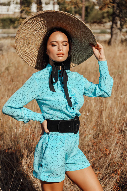 fashion summer outdoor photo of beautiful sexy woman with dark hair in elegant dress with luxurious straw hat and bag posing in field with dry grass - Фото, зображення