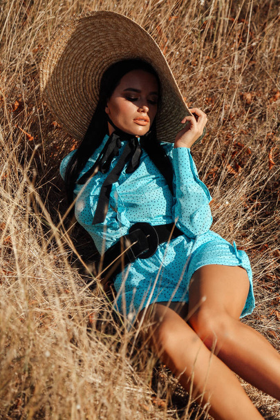 fashion summer outdoor photo of beautiful sexy woman with dark hair in elegant dress with luxurious straw hat and bag posing in field with dry grass - Фото, зображення