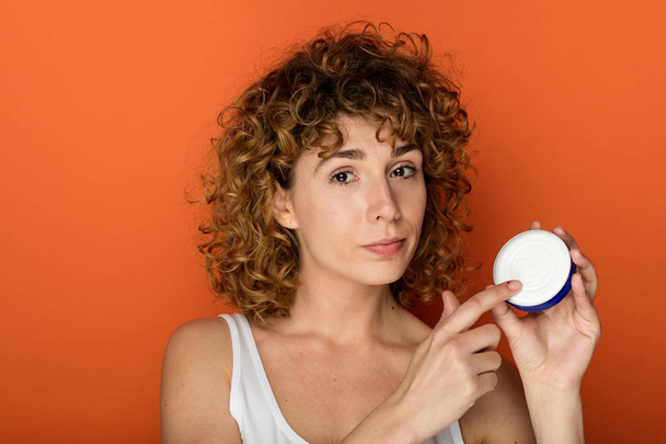 young curly woman holding a small box of skin cream on orange background - Valokuva, kuva