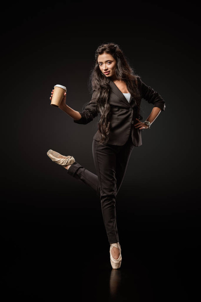 attractive young businesswoman in formal wear and ballet shoes with coffee to go on black background  - Foto, Imagen