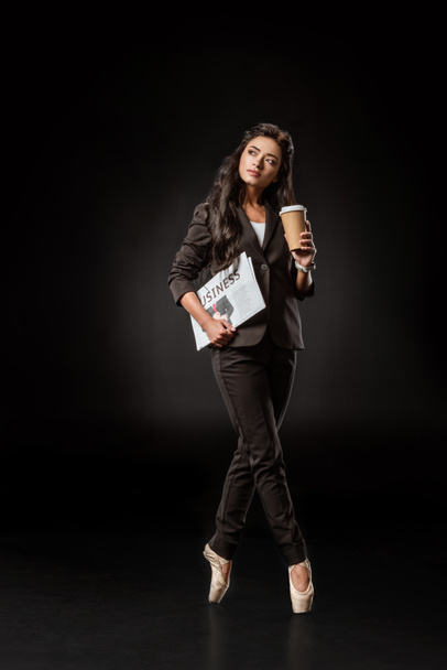pensive businesswoman in ballet shoes with newspaper and coffee to go on black backdrop - Fotó, kép