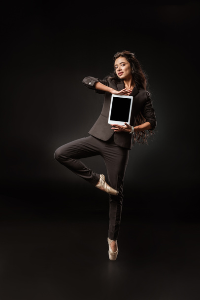 schöne Geschäftsfrau in Anzug und Ballettschuhen zeigt Tablet mit leerem Bildschirm auf schwarzem Hintergrund - Foto, Bild