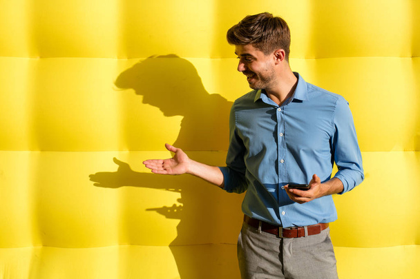 elegant smart casual guy with cellphone presenting an offer on his side, standing on a yellow background - Foto, Imagen