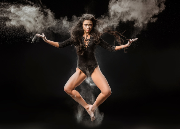 beautiful ballerina in black bodysuit jumping on dark background with talc powder around - Photo, Image
