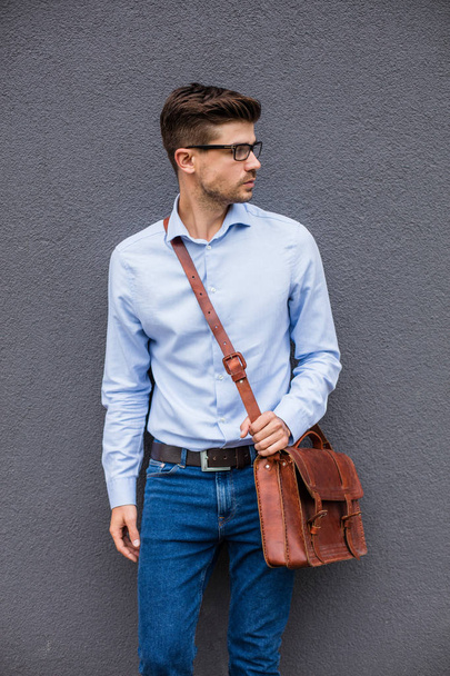 smart and cool. smart casual man with eyeglasses and leather bag standing on gray background, wall, confident and relaxed - 写真・画像