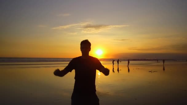 Lassú mozgás szoros fel Silhouette izgatott fiatal férfi ujjongott emelt karokkal. Az emberek a mágikus narancssárga naplemente álmodozó homokos strandon. Tengerparton, kezében az izgalomtól, napkeltekor outstretching boldog srác - Felvétel, videó